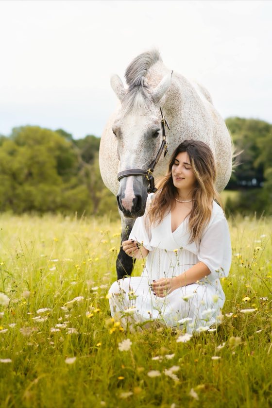 Cavalière en robe blanche assise dans les marguerites auprès de son cheval gris
